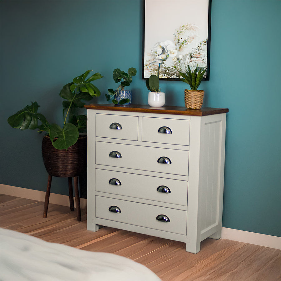 The front of the Alton 5 Drawer Pine Tallboy. There is a free standing potted plant next to it. There are three potted plants on top.