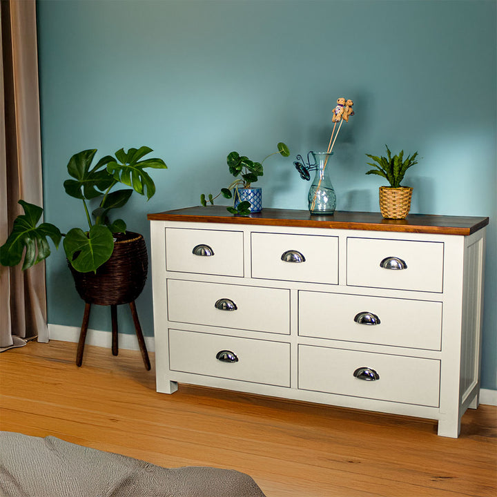 The front of the Alton 7 Drawer Pine Lowboy. There are two blue glass ornaments on top with a pot of white flowers in between. There is a free standing potted plant next to the lowboy.