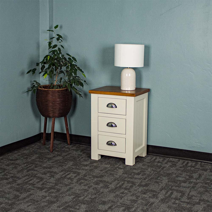 The Alton 3 Drawer Pine Bedside Cabinet with a Rimu stained top. There is a lamp on top and a free standing potted plant next to it.