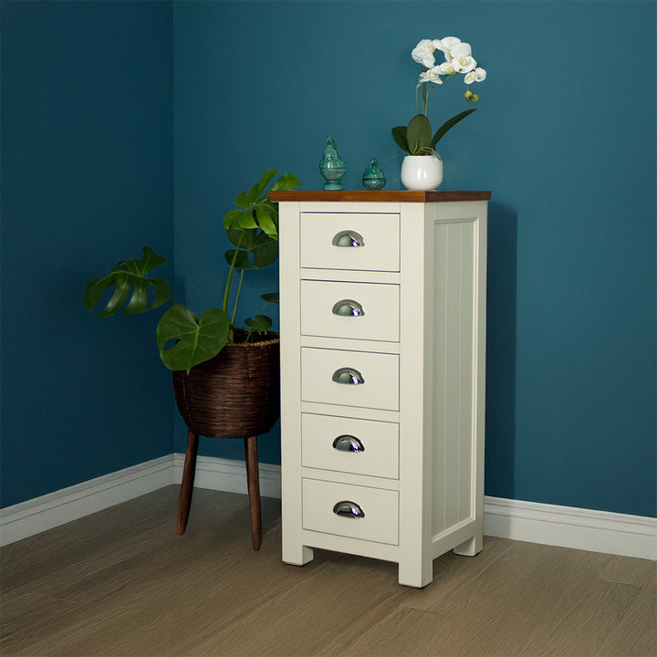 The front of the Alton 5 Drawer Lingerie Chest. There is a free standing potted plant next to it. There are two blue glass ornaments and a pot of white flowers on top.