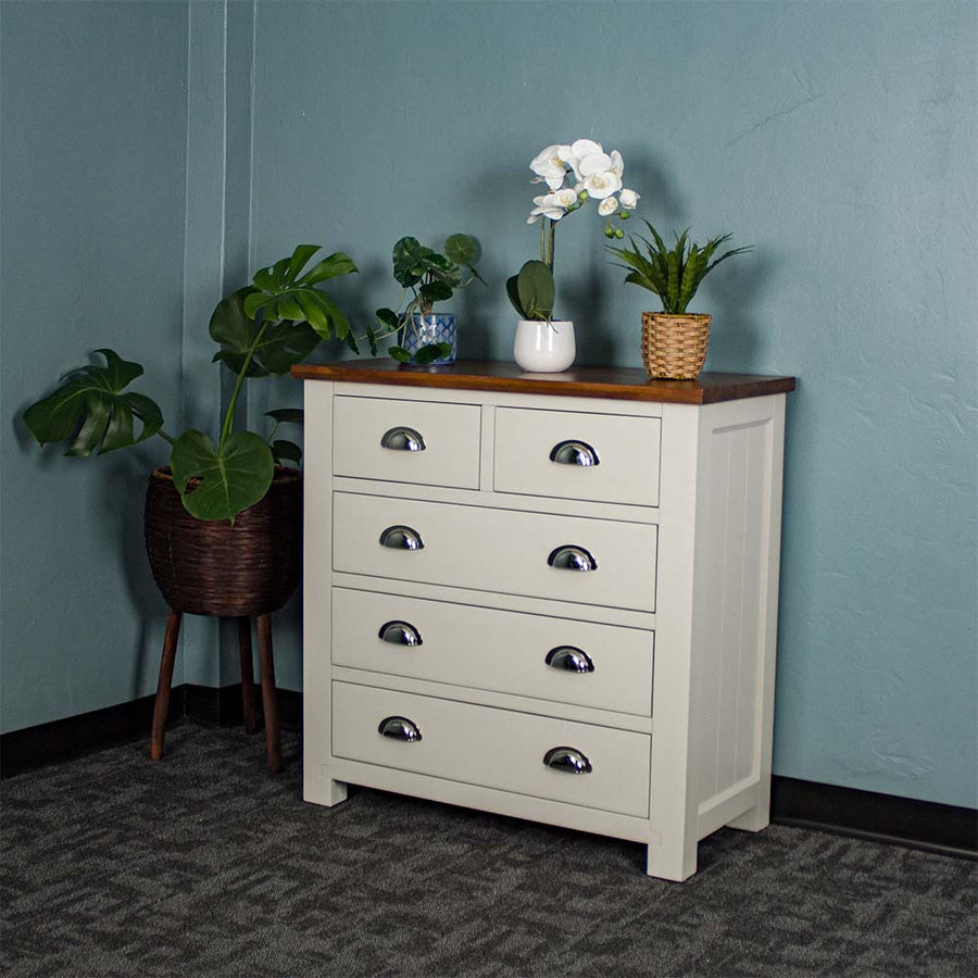 The front of the Alton 5 Drawer Pine Tallboy. There is a free standing potted plant next to it. There are three potted plants on top.