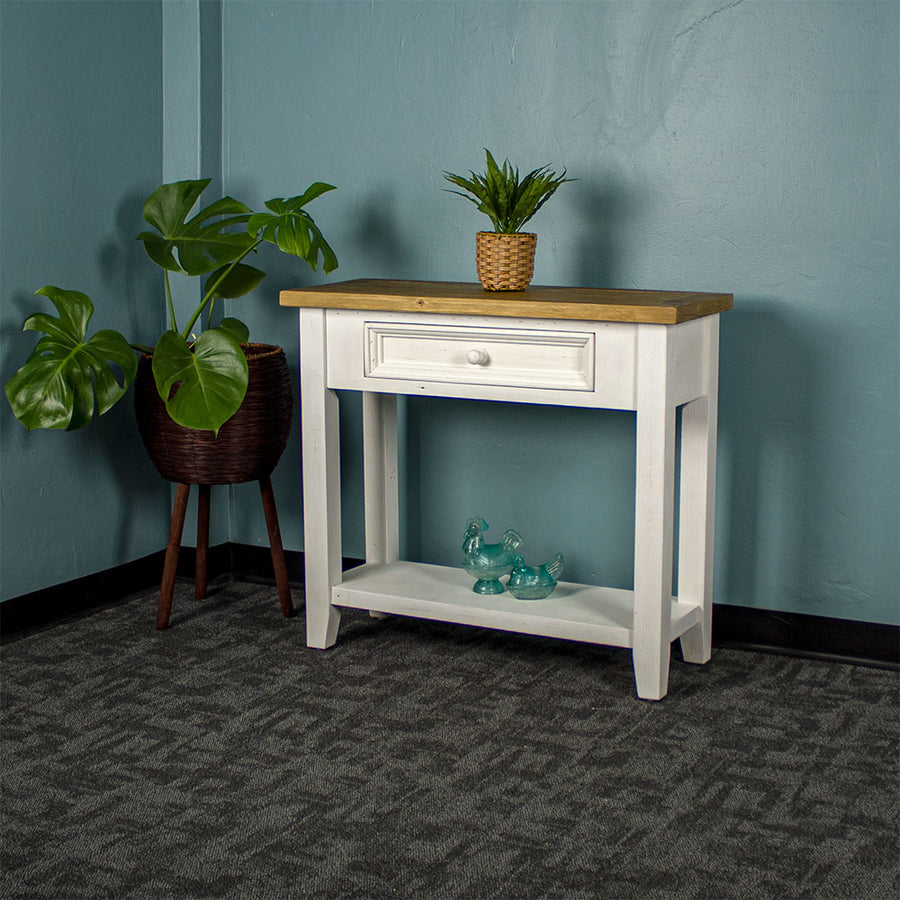 Front view of the Byron Recycled Pine Hall Table. There is a standing potted plant to the side, a small potted plant on top and two blue glass ornaments on the lower shelf.