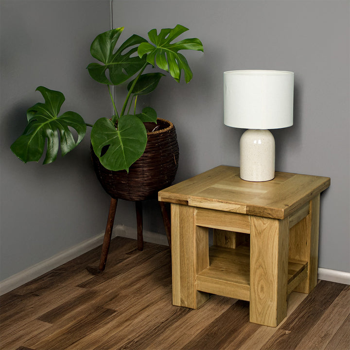 An overall view of the Camden Oak Lamp Table. There is a lamp on top and a free standing potted plant to the side.