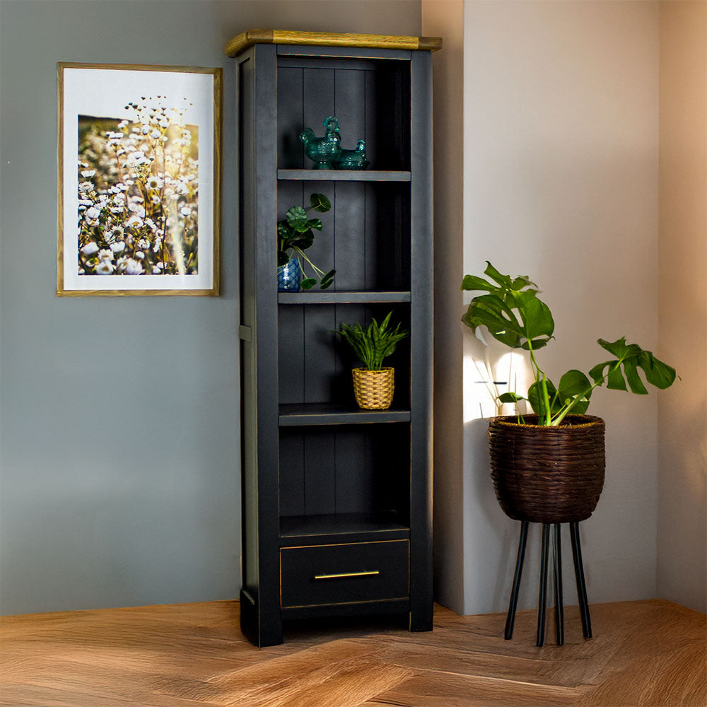 The front of the Cascais Tall Black Bookcase. There is a framed photo of flowers on the wall on the left and a tall, free standing potted plant on the right on the ground. There are two blue glass ornaments on the top shelf, a potted plant on the second shelf, and a third on the bottom shelf.