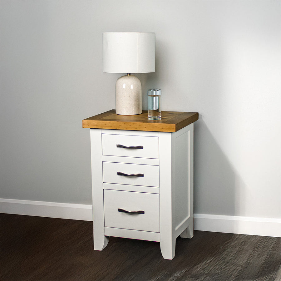 The front of the Felixstowe Pine Bedside Cabinet (White). There is a lamp and a glass of water on top.