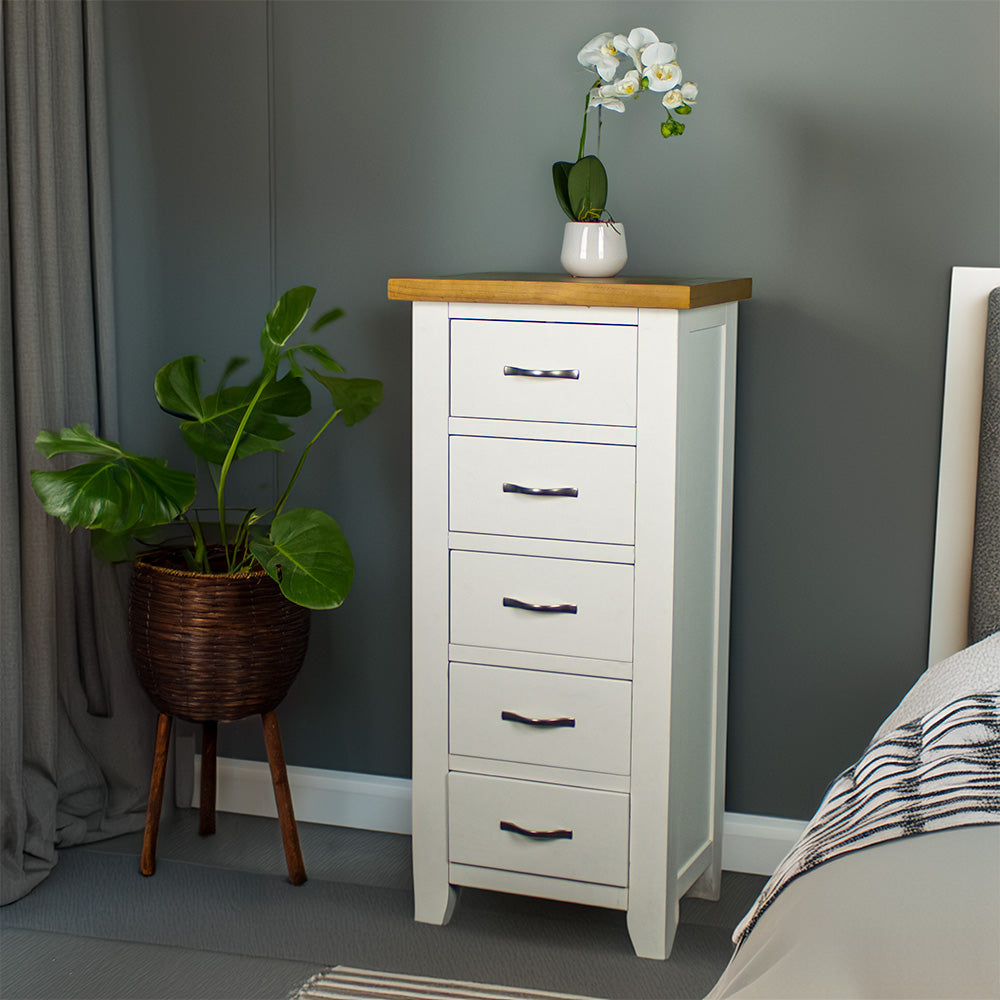 The front of the Felixstowe 5 Drawer Pine Lingerie Chest. There is a small pot of white flowers on top. There is a free standing potted plant next to the unit.