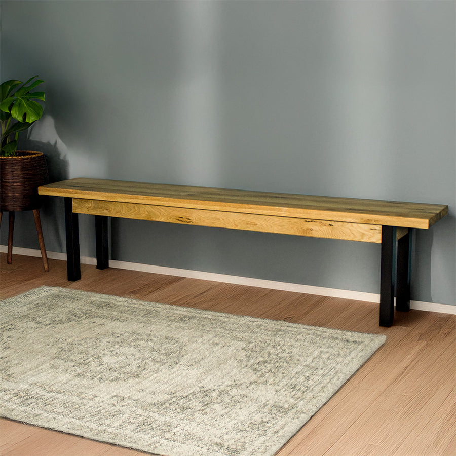 The front of the Golden Gate Long Oak Bench. There is a large gray rug in front and a potted plant next to the bench.