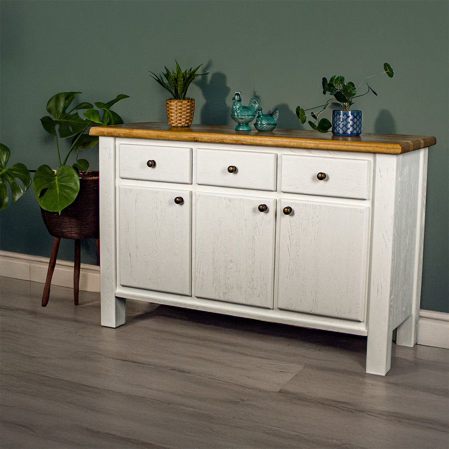 The front of the Loire Two-Tone Medium Oak Buffet. There are two potted plants on top with two blue glass ornaments in between. There is a free standing potted plant to the left of the sideboard.