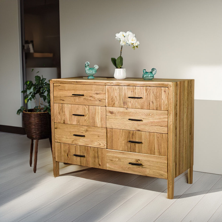 The front of the Ormond 8 Drawer Oak Tallboy. There are two blue glass ornaments on top with a pot of white flowers in between. There is a free standing potted plant next to it.