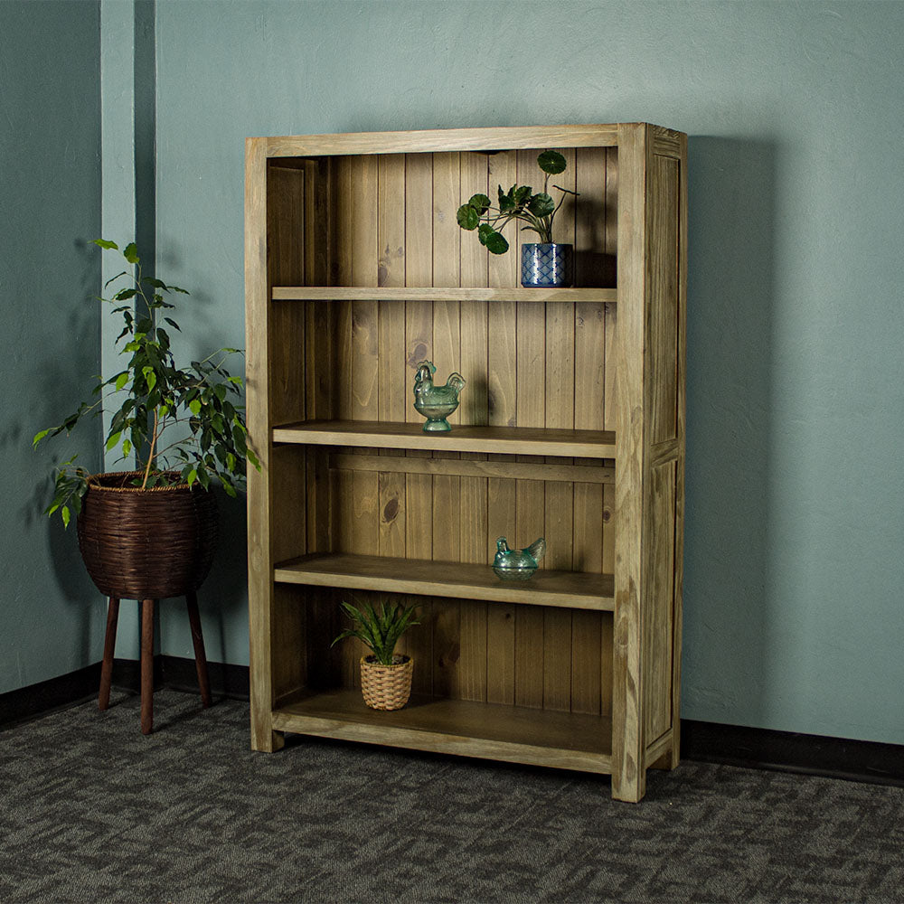 The front of the Vancouver 1.5 m Pine Bookcase. There is a potted plant on the top shelf, a blue glass ornament on the second shelf, a blue glass ornament on the third shelf and a potted plant on the lowest shelf. There is a free standing potted plant next to the bookshelf.