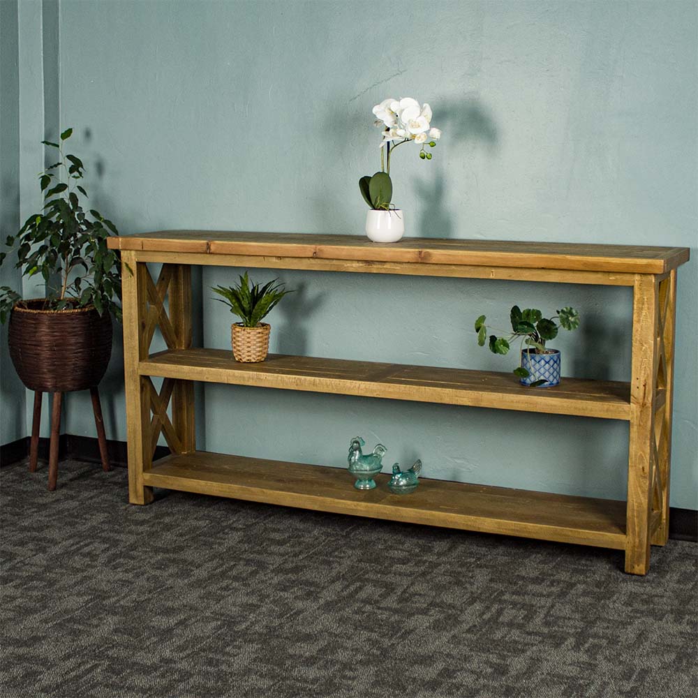 The front of the Ventura Recycled Pine Large Hall Table. There is a free standing potted plant next to it. There is a pot of white flowers on top. There are two equidistant potted plants on the middle shelf. There are two blue glass ornaments on the bottom shelf.