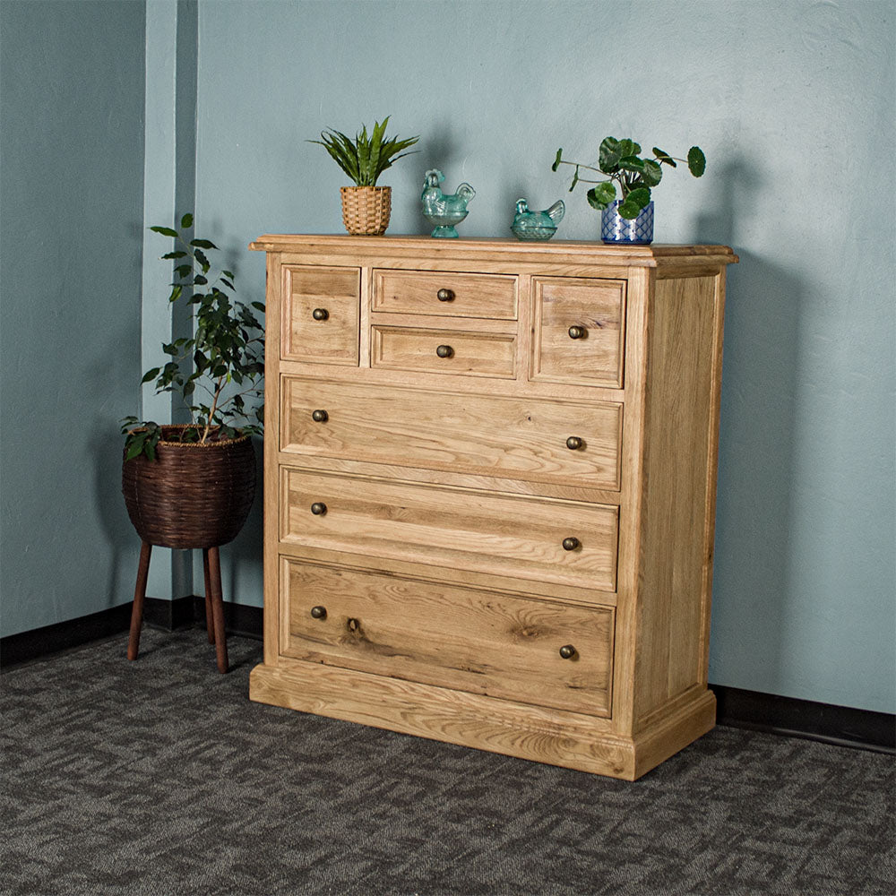 The front of the Versailles Oak 7 Drawer Chest. There are two potted plants on top, as well as two blue glass ornaments. There is a free standing potted plant next to it as well.