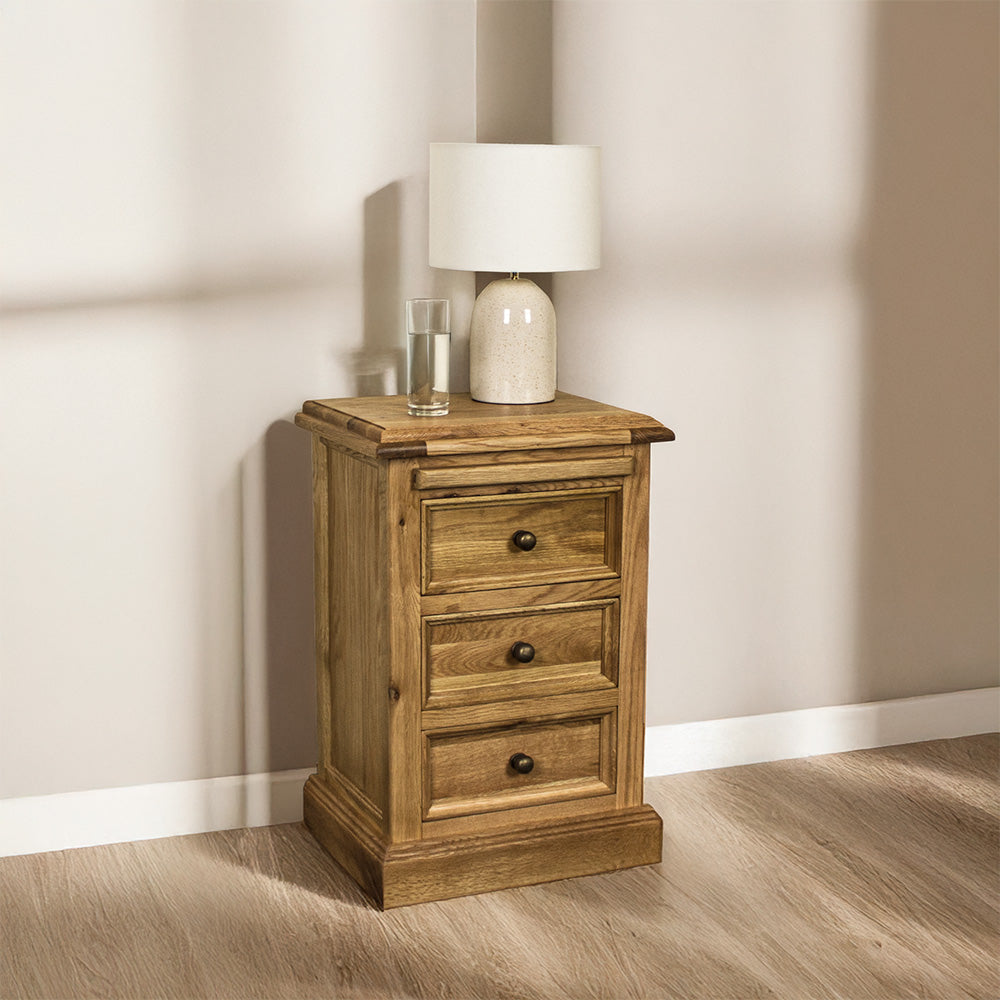 The front of the Versailles Oak Bedside Cabinet. There is a lamp and a glass of water on the top.