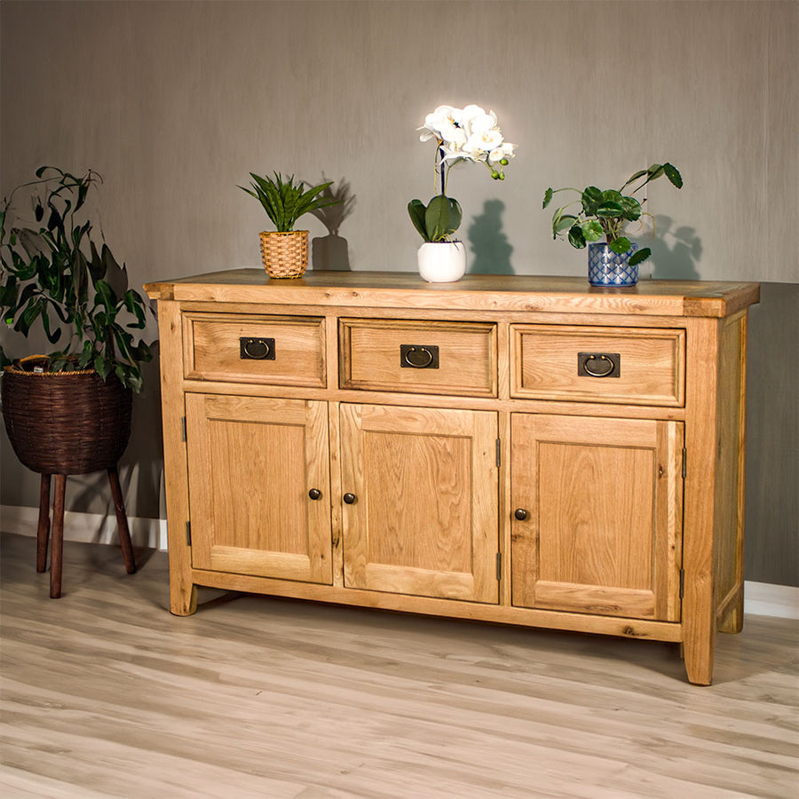 The front of the Yes 3 Door 3 Drawer Oak Buffet. There are three potted plants on top of the unit and a free standing potted plant next to it.