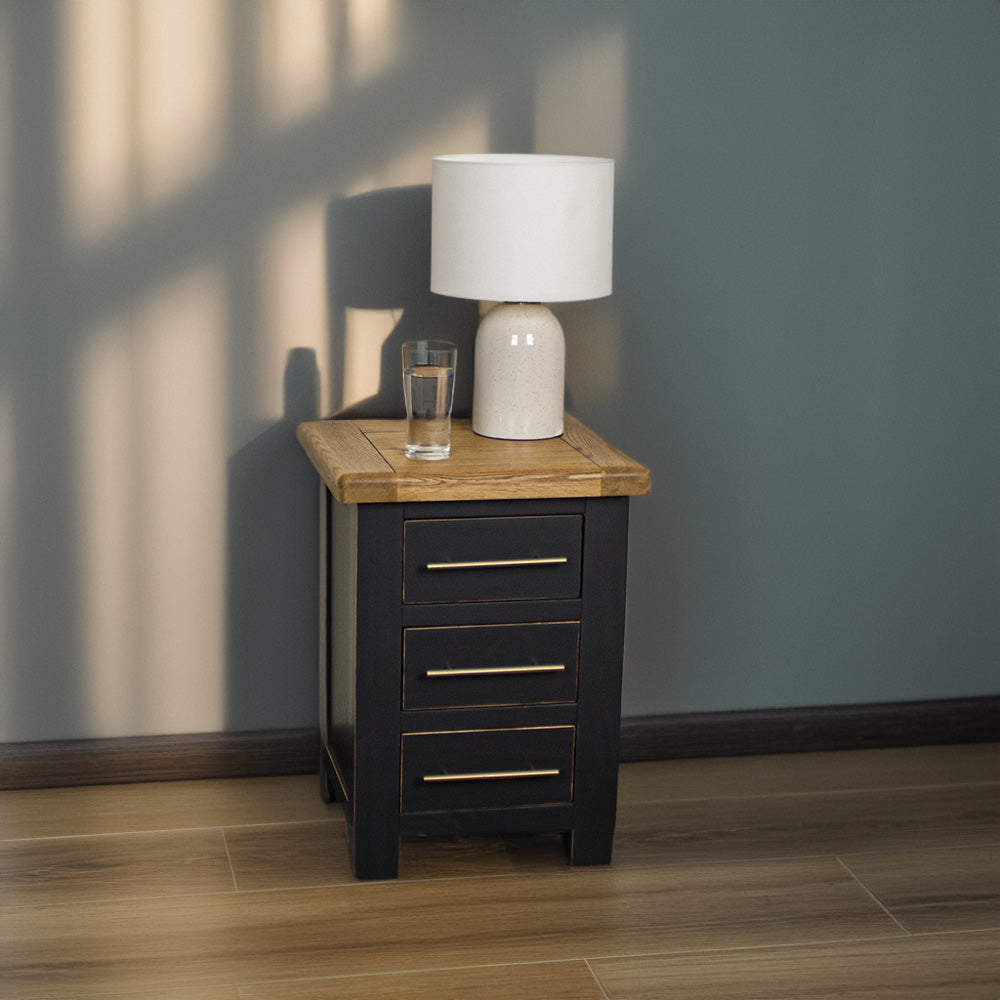 The front of the Cascais 3 Drawer Black Bedside Table. There is a glass of water and a lamp on top.