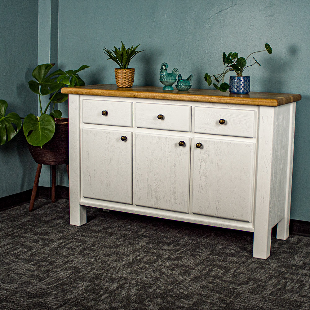 The front of the Loire Two-Tone Medium Oak Buffet. There are two potted plants on top with two blue glass ornaments in between. There is a free standing potted plant to the left of the sideboard.