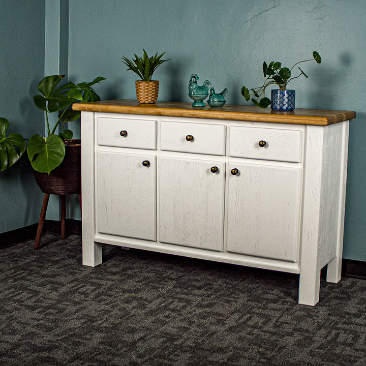 The front of the Loire Two-Tone Medium Oak Buffet. There are two potted plants on top with two blue glass ornaments in between. There is a free standing potted plant to the left of the sideboard.