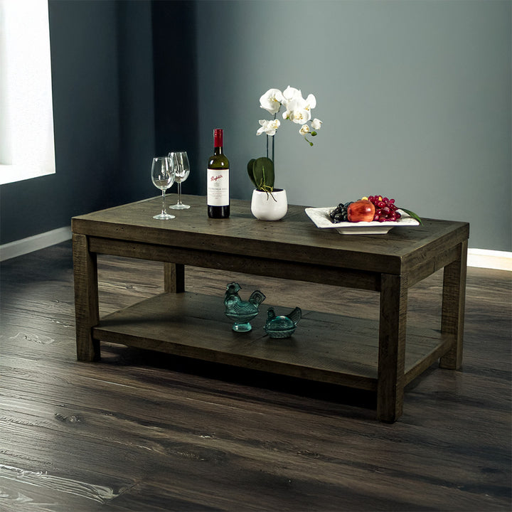 Front view of the brown Stonemill Recycled Pine Coffee Table. There are two wine glasses, a glass bottle, a potted plant and a platter of fruit on the top. There are two blue glass ornaments on the bottom shelf. There is also text in the bottom right corner that says "100% solid recycled pine" and "Environmentally friendly"