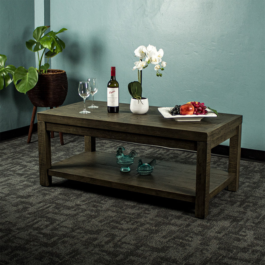 Front view of the brown Stonemill Recycled Pine Coffee Table. There are two wine glasses, a glass bottle, a potted plant and a platter of fruit on the top. There are two blue glass ornaments on the bottom shelf. There is also text in the bottom right corner that says "100% solid recycled pine" and "Environmentally friendly"