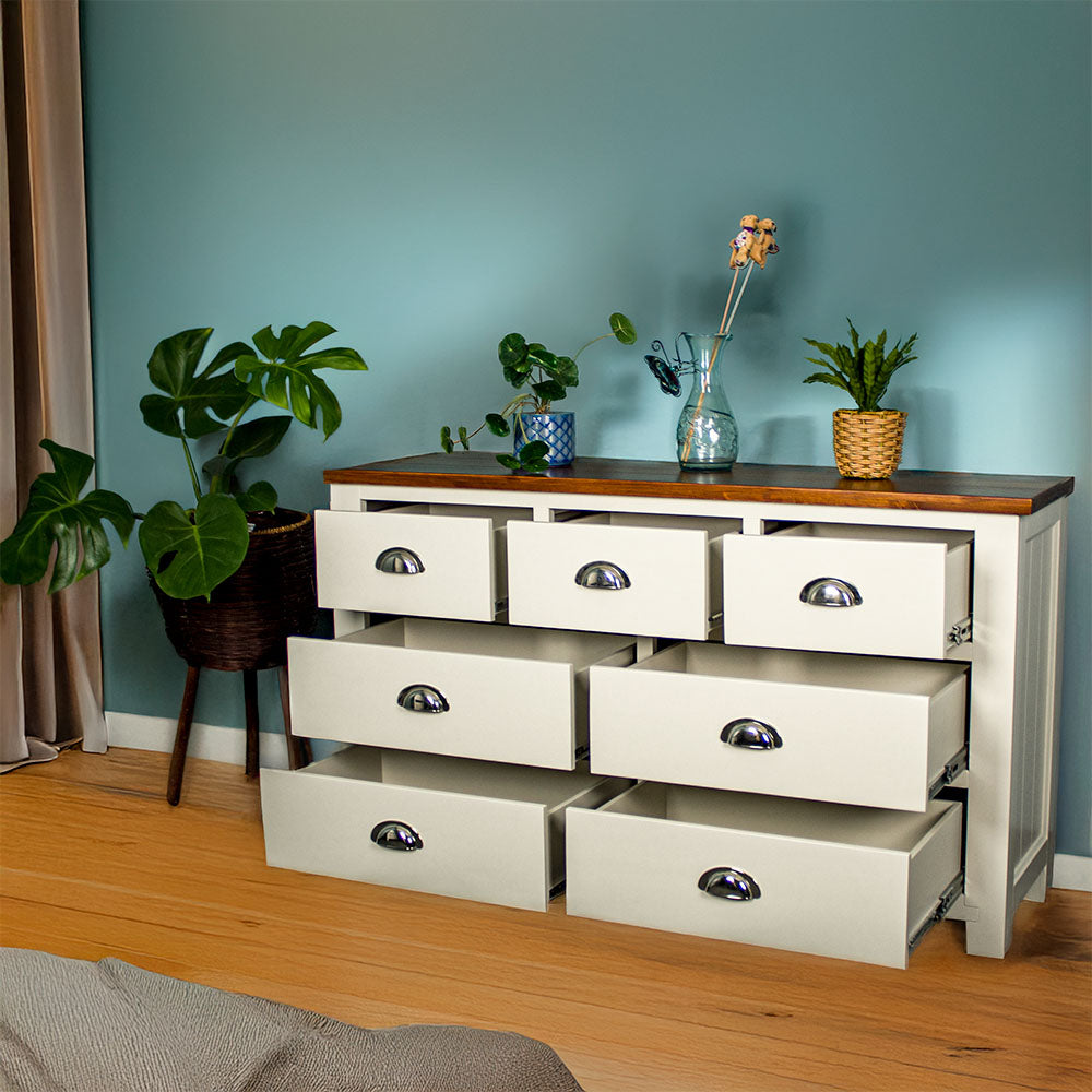 The front of the Alton 7 Drawer Pine Lowboy with its drawers open. There are two blue glass ornaments on top with a pot of white flowers in between. There is a free standing potted plant next to the lowboy.