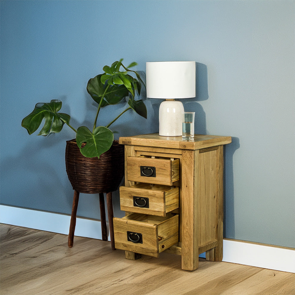 The front of the Amstel Oak Bedside Table with its drawers open. There is a free standing potted plant next to it. There is a lamp and a glass of water on top.