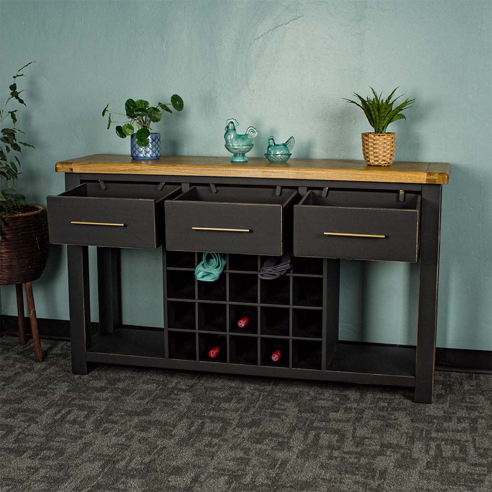 The front of the Cascais Large Buffet with Wine Rack with its drawers open. There is a tall potted plant next to the unit. There are two small potted plants on top in between two blue glass ornaments. There are three wine bottles in the wine rack and two towels in the top.