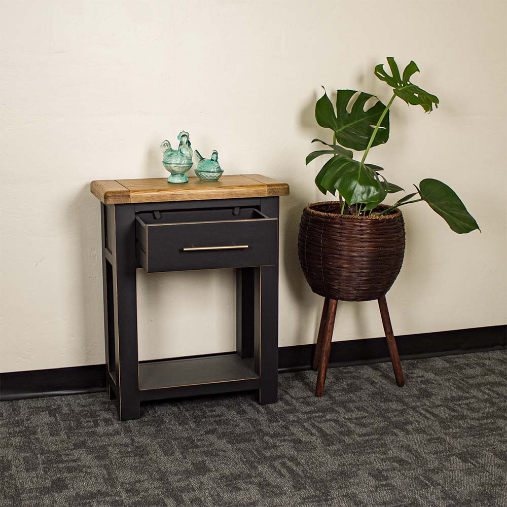 An overall view of the Cascais Black Hall Table, with its drawer open. There are two blue glass ornaments on top with a tall, free standing potted plant next to it.