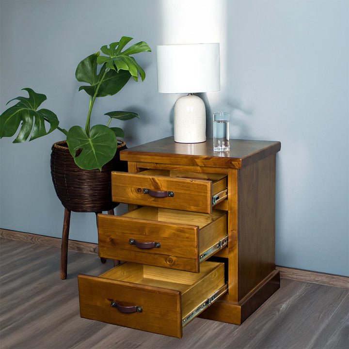 The front of the Jamaica Bedside Cabinet with its drawers open. There is a lamp and a glass of water on top. There is a potted plant next to the bedside table.