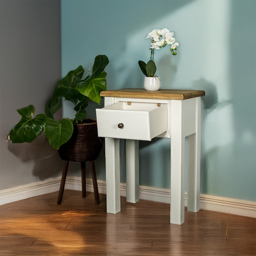 The front of the Loire Oak Console Table with its drawer open. There is a pot of white flowers on top and a free standing potted plant to the right.