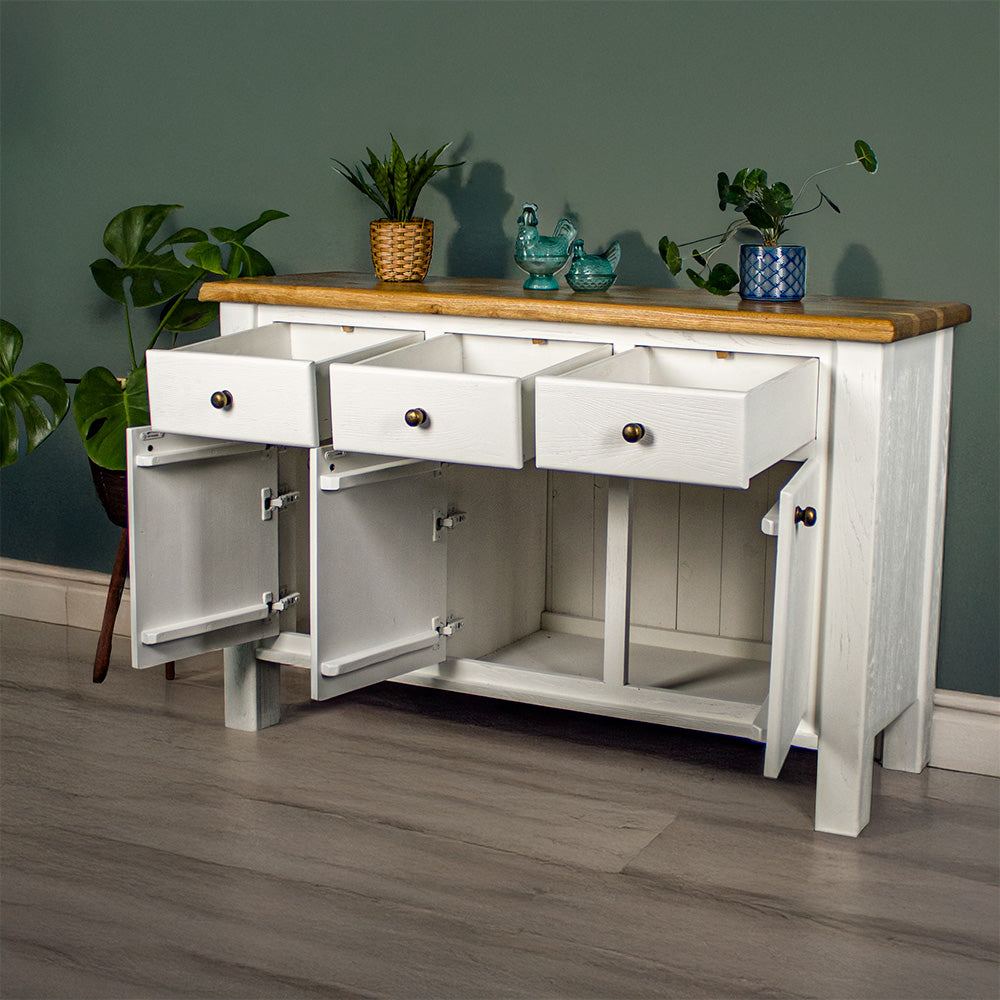 The front of the Loire Two-Tone Medium Oak Buffet with its drawers and doors open. There are two potted plants on top with two blue glass ornaments in between. There is a free standing potted plant to the left of the sideboard.