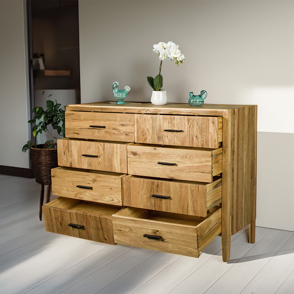 The front of the Ormond 8 Drawer Oak Tallboy with its drawers open. There are two blue glass ornaments on top with a pot of white flowers in between.