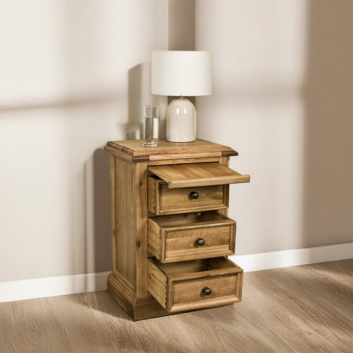 The Versailles Oak Bedside Cabinet with its drawers open. There is a lamp and a glass of water on top.