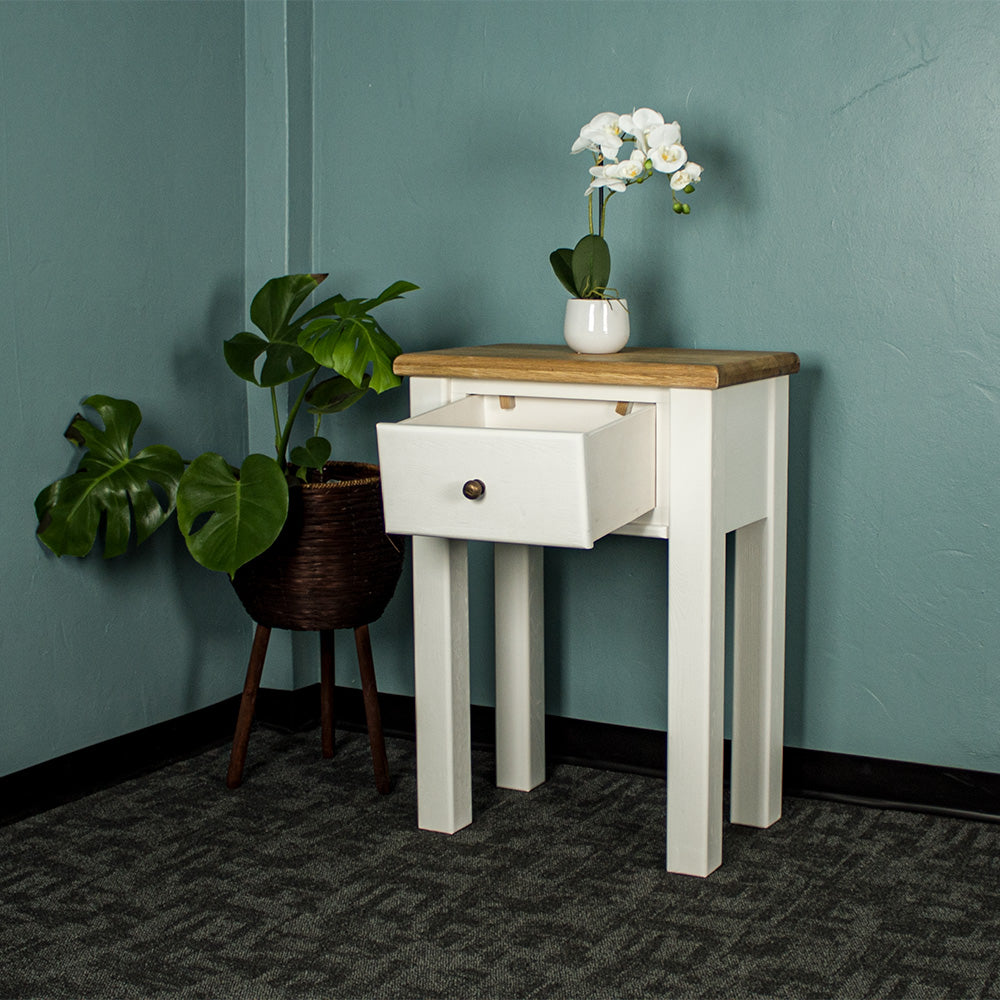 The front of the Loire Oak Console Table with its drawer open. There is a pot of white flowers on top and a free standing potted plant to the right.