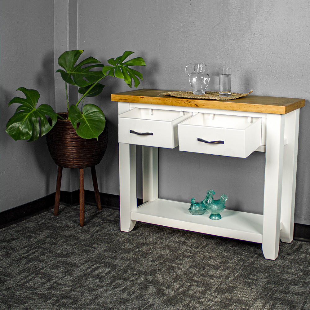 The front of the Felixstowe Large Pine Hall Table with its drawers open. There are two blue glass ornaments on the lower shelf, and a glass water pitcher and glass of water on a rattan leaf on top of the console table. There is a free standing potted plant next to the hallway table.