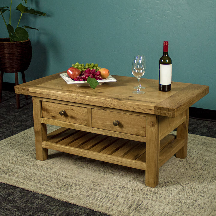 The front of the Farmhouse Coffee Table, which is sitting on a gray rug. There is a free standing potted plant in the back. There is a fruit platter, two wine glasses and a wine bottle on top.