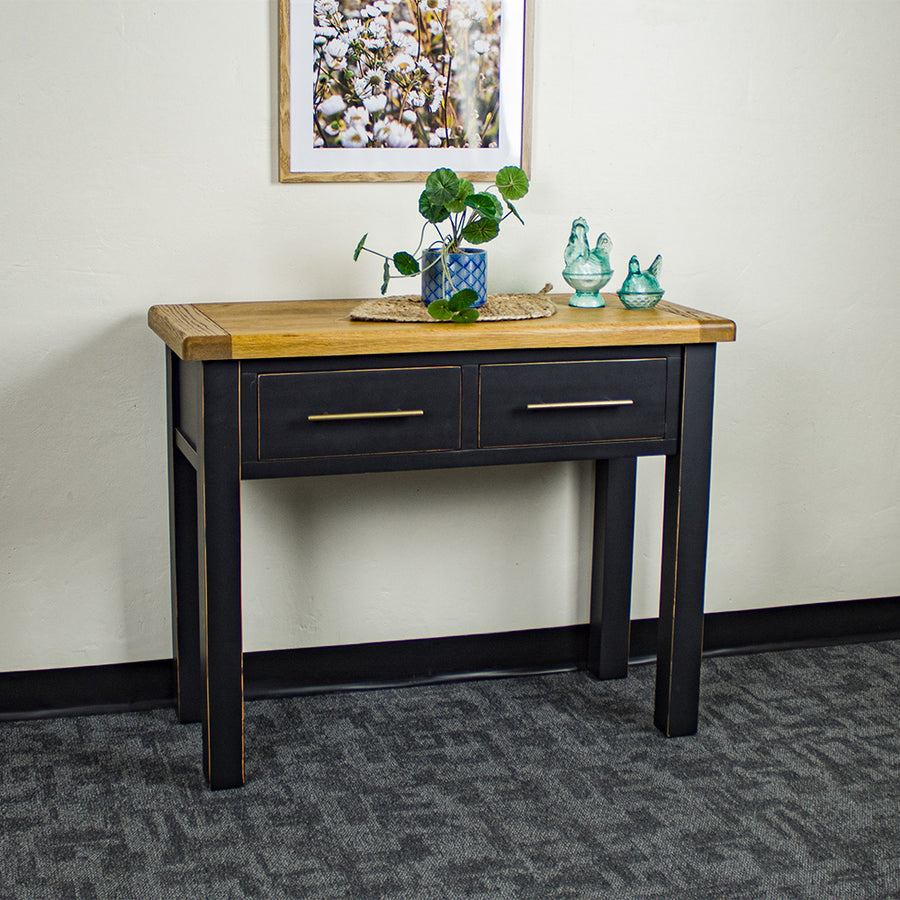 The front of the Cascais 2-Drawer Hall Table. There is a potted plant on top of a rattan leaf, in the middle on the top. There are two blue glass ornaments on top. There is a picture of flowers on the wall.