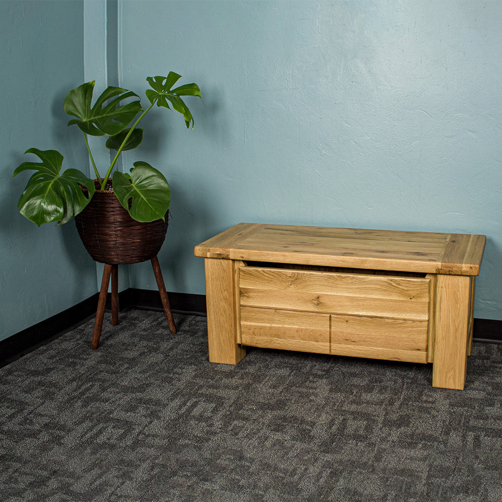 An overall view of the Camden Oak Blanket Box. There is a free standing potted plant next to it.