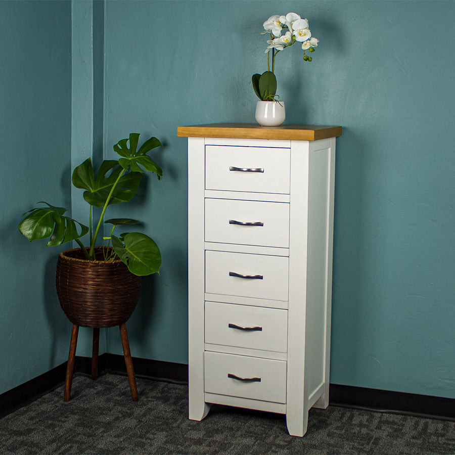 The front of the Felixstowe 5 Drawer Pine Lingerie Chest. There is a small pot of white flowers on top. There is a free standing potted plant next to the unit.